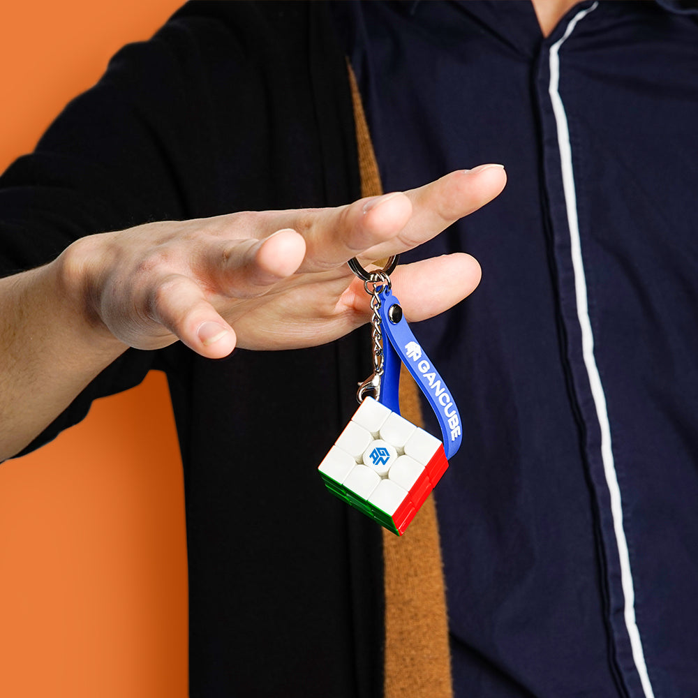 Hand holding a GanCube 3x3 keychain puzzle with blue strap against orange background