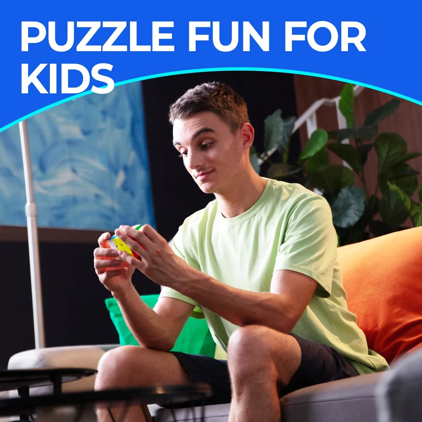 Young man in casual clothes solving a colorful Rubik's Cube on a grey sofa with vibrant cushions