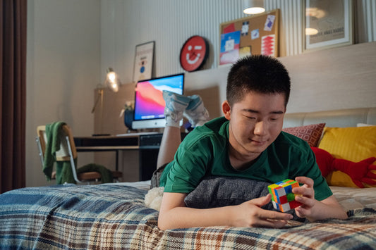 Boy lying on bed solving a colorful Rubik's Cube in a cozy bedroom with computer and decor