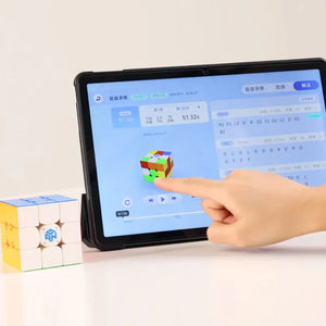 Hand interacting with tablet showing GAN smart cube app beside a solved GAN speedcube on white surface